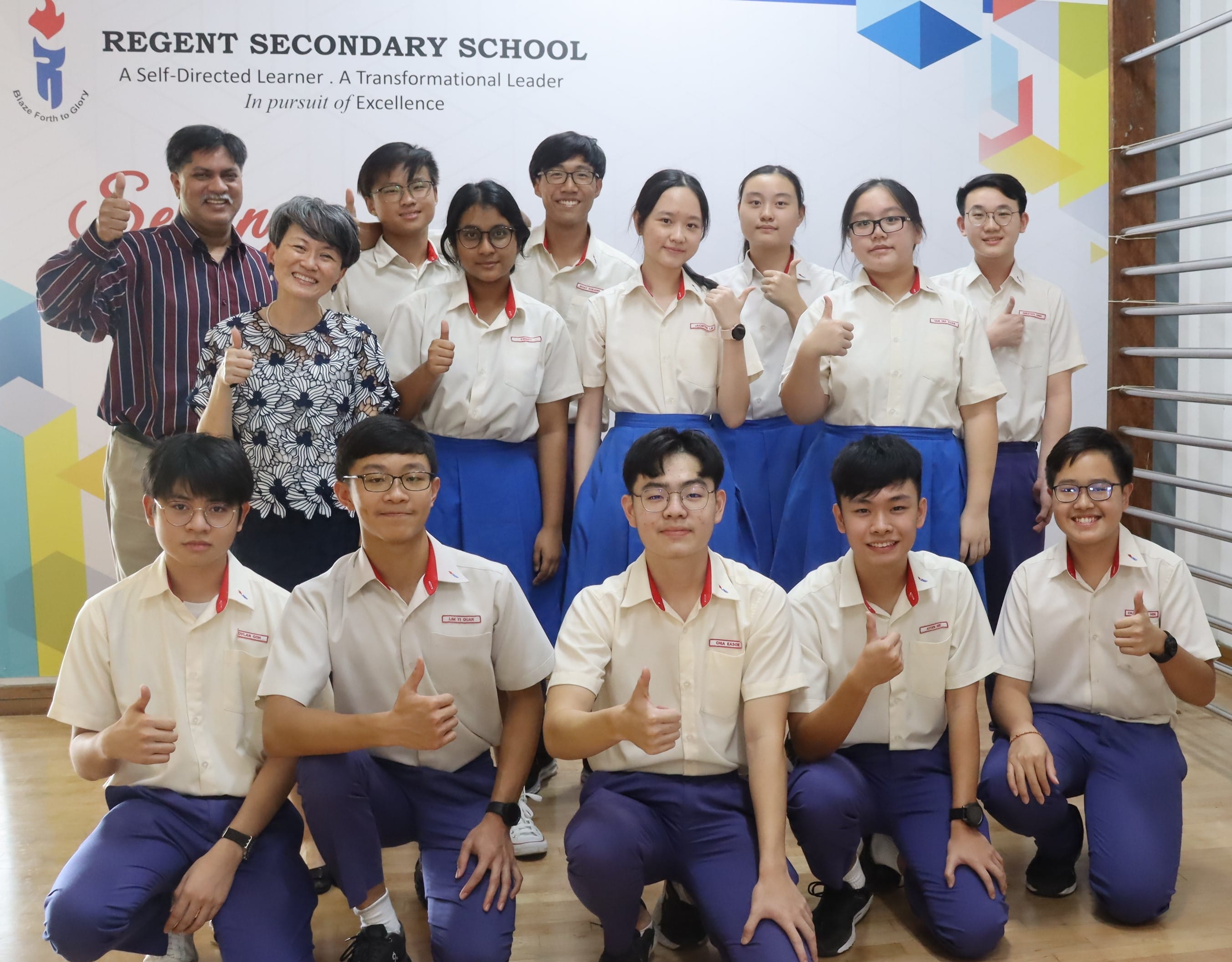 Group of students in uniform and two teachers posing with thumbs up in front of Regent Secondary School banner.